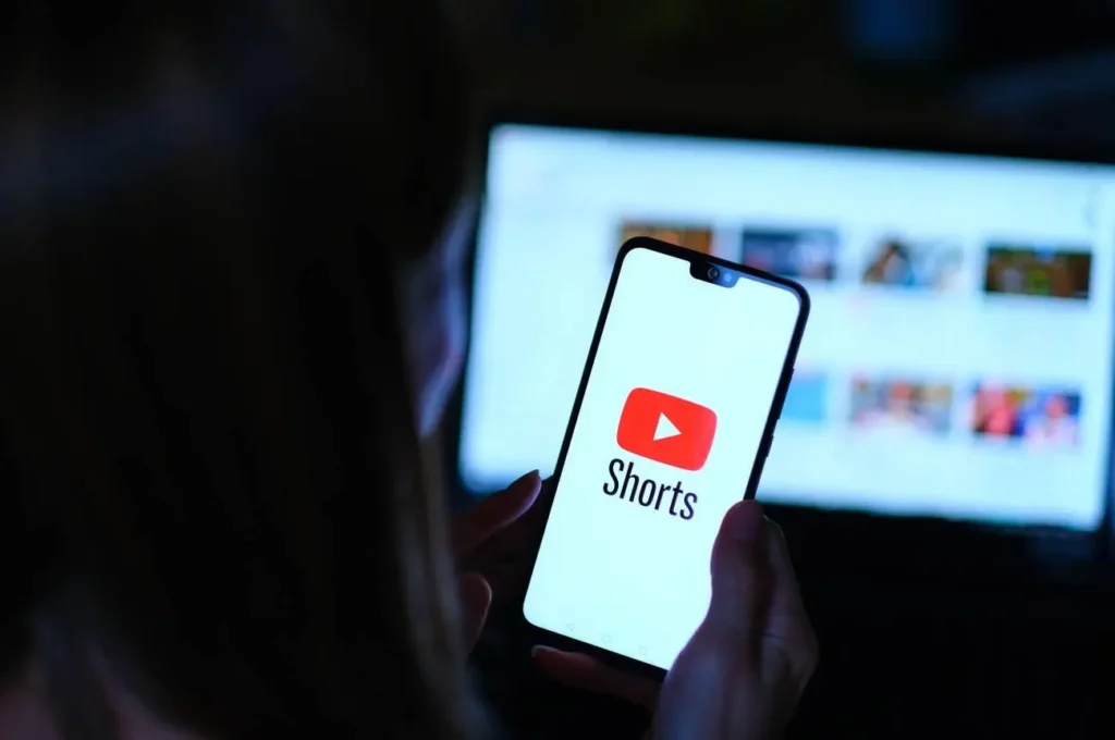 Persona sosteniendo un teléfono móvil con la pantalla mostrando el logotipo de YouTube Shorts, mientras una computadora portátil está abierta en el fondo, parcialmente desenfocad